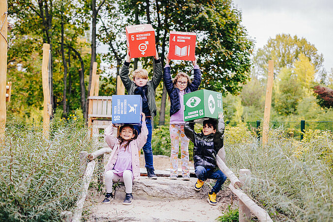 Kinder auf einem Spielplatz halten große Würfel mit Nachhaltigkeitszielen in die Luft.