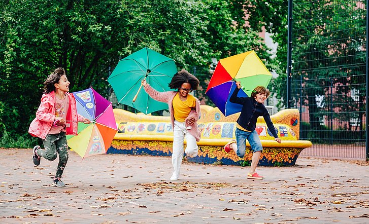 Kinder rennen mit offenen Regenschirmen fröhlich über einen Spielplatz