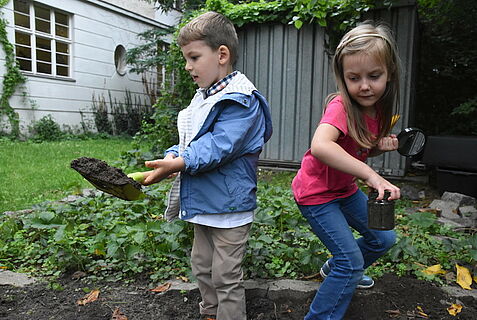 Ein Junge grabt Erde in einem Beet um. Ein Mädchen hält ein mit Erde befülltes Glas und eine Lupe in ihren Händen.