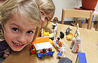 Zwei Mädchen sitzen am Tisch mit Spielzeugfiguren und lachen aktiv in die Kamera 