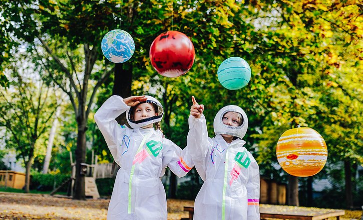 Zwei als Astronauten verkleidete Kinder zeigen auf bunte Planeten