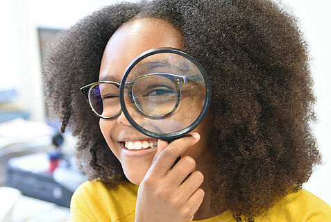 Ein Mädchen mit Brille schaut durch eine Lupe in die Kamera.