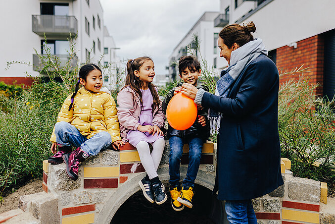 Eine Kita-Fachkraft hält einen Luftballon in der Hand, drei Kita-Kinder sitzen dahinter auf einer Mauer