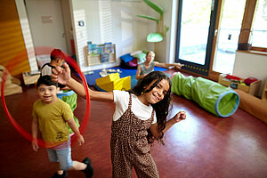 Im Vordergrund schwingt ein Mädchen einen Reifen an ihrem rechten Handgelenk und lächelt dabei in die Kamer. Im Hintergrund sieht man zwei weitere Kinder in Bewegung und eine Fachkraft, die auf dem Boden sitzt.