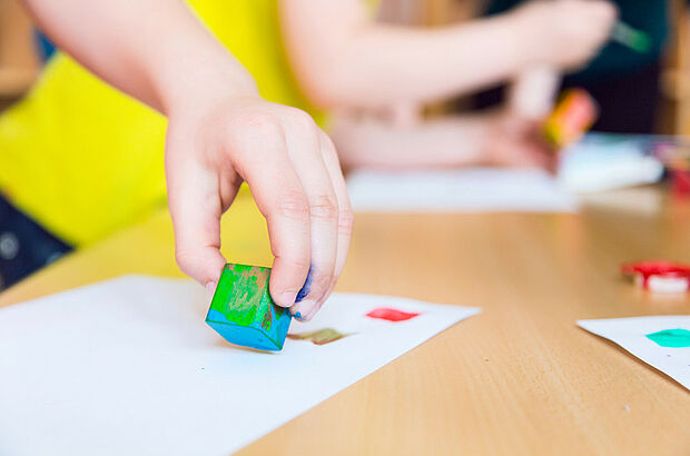 Kinderhand stempelt grünen Baustein mit Farbe auf Papier.