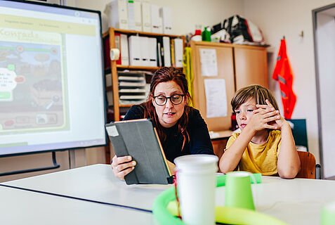 Lehrerin mit Schüler in Klassenzimmer, Lehrerin hält Tabletin der Hand, im Hintergrund sieht man ein aktives Smartboard