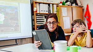 Lehrerin mit Schüler in Klassenzimmer, Lehrerin hält Tabletin der Hand, im Hintergrund sieht man ein aktives Smartboard