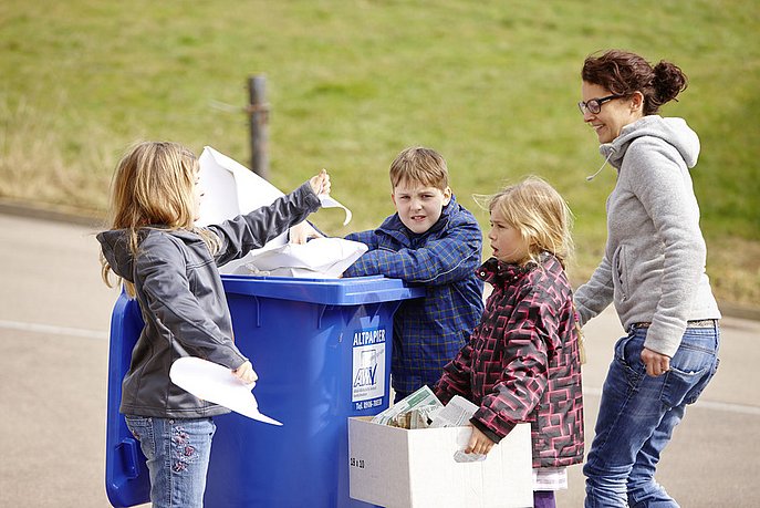Drei Kinder und eine Frau stehen mit einem Karton und verschiedenen Papiersorten an einer Papiertonne.