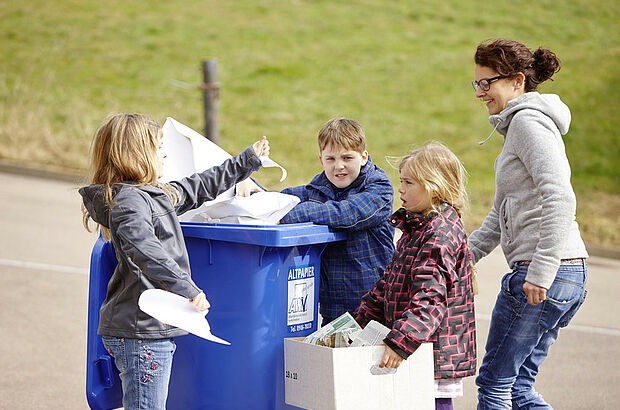 Drei Kinder und eine Frau stehen mit einem Karton und verschiedenen Papiersorten an einer Papiertonne.