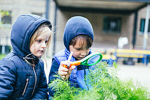 Zwei Kinder betrachten draußen mit einer Lupe eine Pflanze.