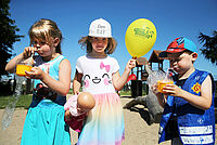 Zwei Kinder pusten durche inen Strohhalm in eine Schale, sodass Blasen entstehen. In ihrer Mitte ein Mädchen mit gelben Luftballon.