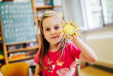 Kind steht vor einer Tafel und hält eine gebastelte Sonne in der Hand.