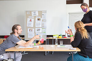 Das Bild zeigt drei Personen bei einer Fortbildungen, die verschiedene Sachen in der Hand haben und miteinander sprechen