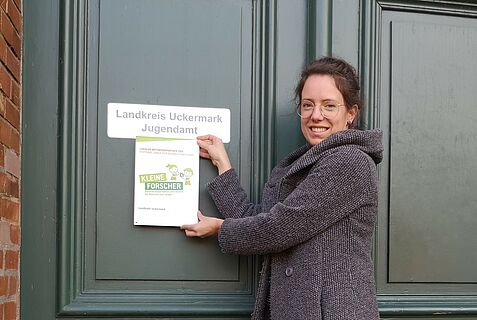 Eine Frau hält eine Plakette "Haus der kleinen Forscher" an eine Tür.