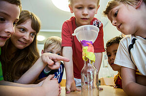 Eine Frau schaut sich gemeinsam mit fünf Kindern eine Konstruktion aus Knete, Flasche und Trichter an