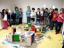 Ein Gruppe Kinder steht um eine Menge gebastelter Häuser in der Mitte des Raumes