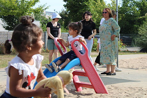 Zwei Kinder spielen im Sommer auf einem Spielplatz. Erwachsene im Hintergrund.