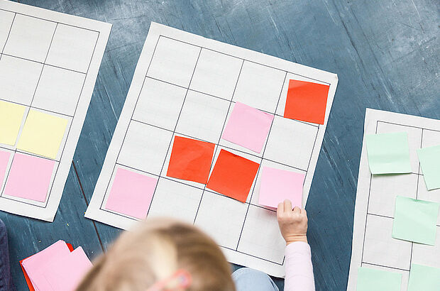 Kinderhand legt quadratische Zettel in ein Raster auf dem Boden