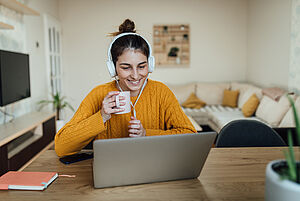 Fröhliche Frau im Pulli mit Kopfhörern vor dem Laptop und Tasse in der Hand