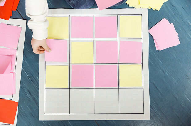 Kinderhand legt quadratische verschiedenfarbige Zettel in ein Raster auf dem Boden