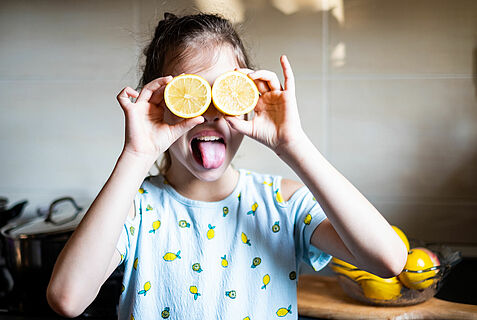 Ein Mädchen hält sich zwei aufgeschnittene Zitronenhälften vor die Augen und streckt die Zunge heraus.