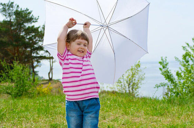 Junges Mädchen steht mit offenem weißem Regenschirm in einer Wiese.