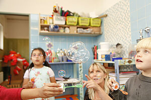Eine Erzieherin schaut mit zwei Kindern Seifenblasen hinterher