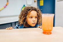 Ein Kind beobachtet staunend ein Glas mit einer orange farbenen Flüssigkeit