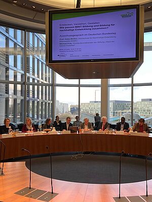 Acht Personen sitzen am Runden Tisch im Bundestag, darüber eine große Präsentationsleinwand