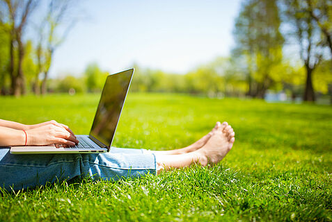 Eine Person sitzt barfuß auf einer Wiese im Park mit einem Laptop auf dem Schoß.