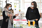 Zwei Frauen, die Maske tragen, stehen hinter einem gebastelten Prototypen und unterhalten sich