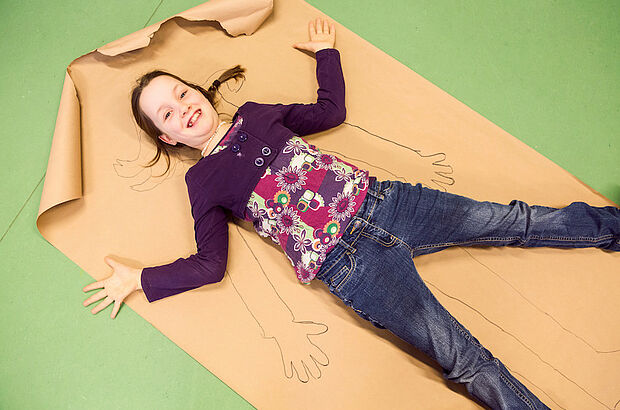 Mädchen liegt auf braunem Papier, auf das sein Umriss gezeichnet wurde.