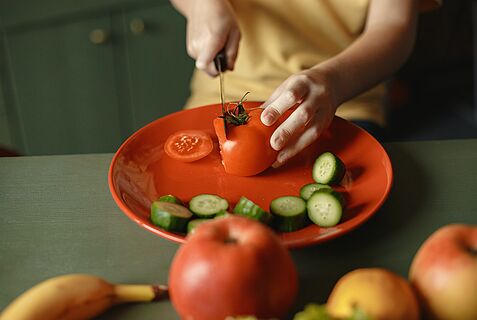 Kinderhände schneiden eine Tomate in Scheiben. Auf dem Teller liegen auch Gurkenstückchen.
