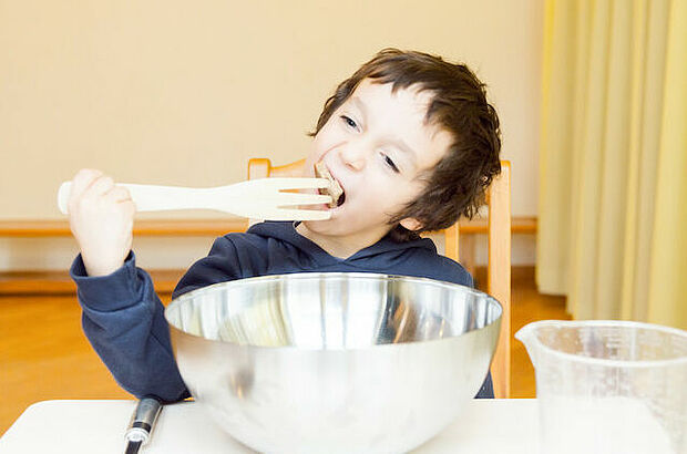 Ein Junge sitzt am Tisch mit übergroßem Geschirr. Er versucht die große Gabel in den Mund zu nehmen.