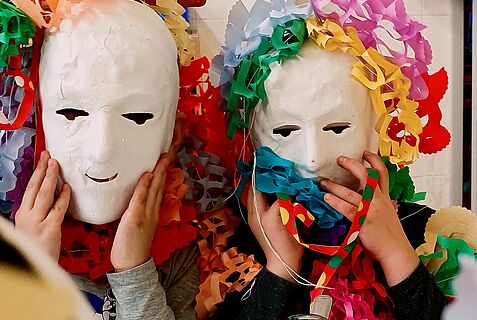 Zwei Kinder tragen weiße Masken und bunte Girlanden