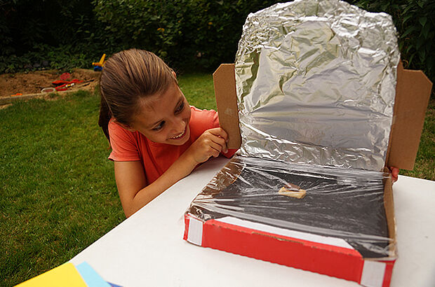 Ein Mädchen blickt auf ein Stück Schokolade in ihrem Solarofen, der aus einem mit Alufolie ausgekleideteten Pizzakarton besteht.