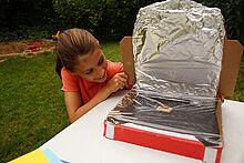 Ein Mädchen blickt auf ein Stück Schokolade in ihrem Solarofen, der aus einem mit Alufolie ausgekleideteten Pizzakarton besteht.