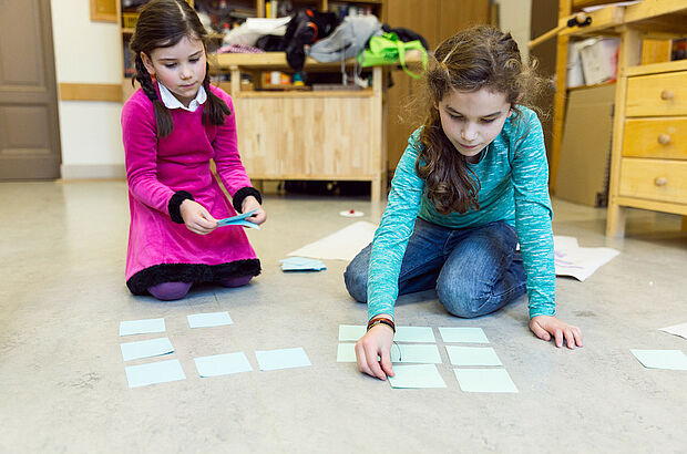 Zwei Mädchen legen quadratische Zettel auf den Boden 