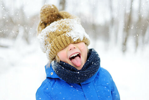 Ein Kind hat eine schneebedeckte Mütze über die Augen gezogen und streckt die Zunge raus.
