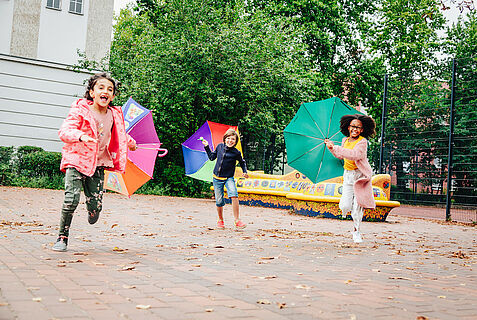 Drei Kinder rennen mit aufgespannten bunten Schirmen der Kamera entgegen.